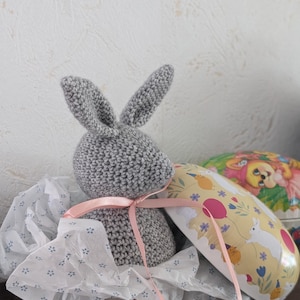 May include: A gray crocheted bunny with long ears, tied with a pink ribbon, rests on patterned tissue paper. Beside it is a yellow Easter egg-shaped container with bunny and chick illustrations. The overall theme is Easter.