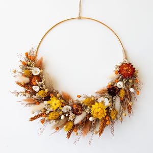 May include: A circular gold metal hoop wreath adorned with an arrangement of dried flowers and grasses in shades of yellow, orange, rust, and cream. The botanical elements are clustered along the lower half of the hoop, suspended by a thin twine.