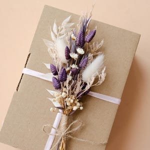 Può includere: Un piccolo bouquet di fiori secchi nei toni del viola, bianco e beige, legato con spago e un nastro rosa, poggia su una scatola di cartone marrone. La composizione include soffici code di coniglio bianche e piccoli fiori bianchi.