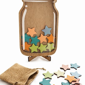 May include: A wooden jar-shaped container with a clear front panel, standing on a small wooden base. Inside the jar are several small, multi-coloured wooden stars in shades of orange, green, and blue. A burlap drawstring bag and loose stars sit nearby.