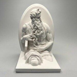 Peut inclure: Une sculpture blanche mate représentant une figure barbue aux cheveux bouclés avec de petites cornes sur le front, vue jusqu'à la taille. Le personnage porte des vêtements drapés, tient une tablette rectangulaire et est placé devant un fond arqué.