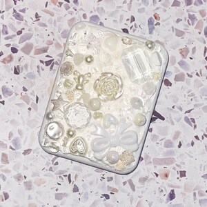 May include: A rectangular white phone case decorated with white and iridescent embellishments. It features pearls, a rose, a bow, stars, and crystal-like accents. The case has a textured, dimensional design.