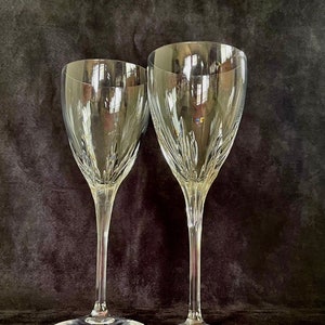 May include: Two clear crystal wine glasses with long stems and wide bases. The glasses have a decorative pattern near the base of the bowl. The background is a dark, textured fabric.