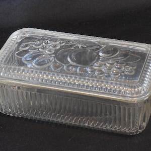 May include: A rectangular clear glass butter dish with a lid. The lid features a molded relief design of grapes and other fruits. The sides of the base and the edges of the lid have a vertical ribbed pattern, providing a textured appearance.