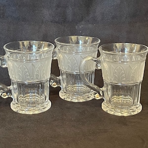 May include: Three clear glass mugs with frosted etched designs. The mugs have a handle and a wide base.