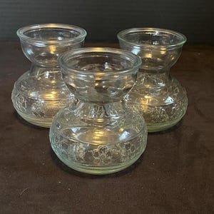 May include: Three clear glass vases with a bulbous base and a flared top. The bases have a floral pattern. The vases are arranged on a dark brown surface.
