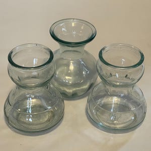 May include: Three clear glass vases with a bulbous base and a narrow neck. The vases are arranged on a white surface. The glass has a slight green tint. The vases are of varying sizes, with the largest in the center.