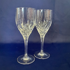 May include: Two clear crystal wine glasses with a decorative pattern on the bowl. The glasses have long stems and round bases. The background is a solid dark blue.