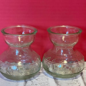 May include: Two clear glass vases with a floral design. The vases have a wide mouth and a narrow neck.