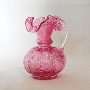 May include: A pink glass pitcher with a ruffled top and a clear glass handle. The pitcher has a textured, bubble-like design.