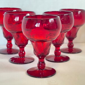 May include: Set of six ruby red wine glasses. Each glass features a rounded bowl, a detailed stem, and a circular base. The glasses are made of transparent glass and are arranged on a white surface.