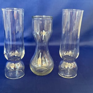 May include: Three clear glass vases of varying shapes. Two are tall and cylindrical, with a bulbous base. The third vase is shorter, with a rounded body and a narrow neck. All are transparent, allowing light to pass through.