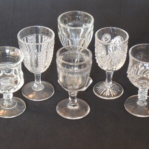 May include: Six clear glass goblets with intricate cut glass designs. The goblets are arranged in a semi-circle on a black surface.