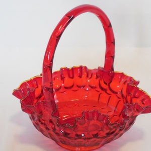 May include: A red glass basket with a ruffled edge and a handle. The basket is made of clear glass with a red tint and has a gold rim.