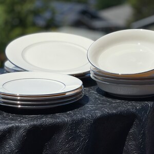 May include: A set of white ceramic dinner plates and bowls with a silver trim. The plates are stacked on top of each other, and the bowls are stacked on top of each other. The plates and bowls are on a black tablecloth.