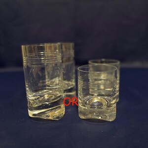 May include: Four clear glass tumblers and lowball glasses are arranged on a dark blue surface. The tall glasses have horizontal etched lines. The lowball glasses have a rounded base and etched lines. The glasses are empty.