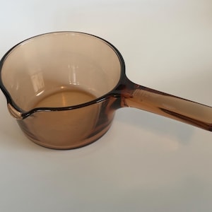 May include: Amber-colored glass saucepan with a long, rectangular handle and a pouring spout. The cookware is made of transparent glass, allowing visibility of the contents. The saucepan is designed for stovetop use.