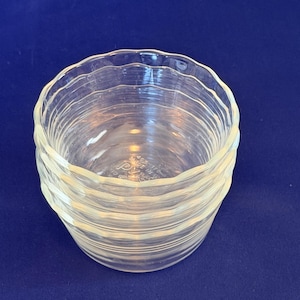 May include: A stack of clear glass bowls with a wavy, ridged design. The bowls are stacked on top of each other, and the edges have a scalloped shape. The bowls are made of clear glass and are sitting on a blue surface.