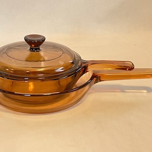 May include: Amber-colored glass cookware set, including a saucepan with a lid and a skillet. The saucepan has a handle and a lid with a knob. The skillet has a handle. The set is made of transparent glass.