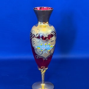 May include: A tall, ruby red glass vase featuring a flared rim and a slender, twisted gold-tone stem on a circular base. The body is decorated with ornate gold filigree patterns and a central hand-painted floral motif in blue, yellow, and white.