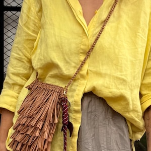 May include: A small tan crocheted crossbody bag featuring long fringe detailing. The bag is attached to a thin braided strap and includes a decorative accessory made of a maroon and white rope cord with a dark crescent-shaped charm attached.