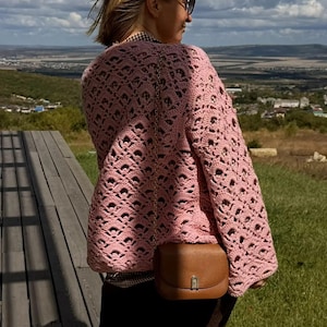May include: A light pink long-sleeved crochet sweater featuring an intricate openwork pattern with a scalloped texture. The garment is paired with a small brown leather crossbody bag that has a gold-toned metal clasp and a thin chain shoulder strap.