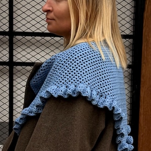 Könnte beinhalten: Ein hellblauer Häkelschal mit gerüschtem Rand. Der Schal wird über einem braunen Mantel getragen. Das Häkelmuster ist sichtbar, und die Rüschen verleihen eine dekorative Note. Der Schal scheint handgefertigt zu sein.