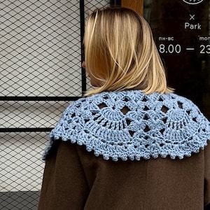 Puede incluir: Un cuello de ganchillo azul claro se usa sobre un abrigo marrón. El cuello presenta un diseño calado detallado con un borde festoneado. La imagen está tomada al aire libre, con un letrero de café visible en el fondo.