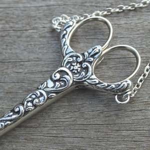 May include: A silver-toned ornate scissors necklace with a chain. The scissors have a detailed floral design.