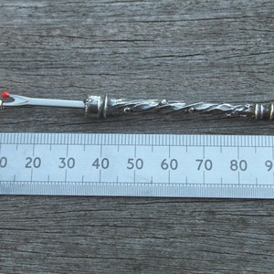 May include: A silver-colored, decorative needle with a red bead at the tip, measuring approximately 9 cm in length. The needle is designed with a twisted, textured body and a small loop at the end.
