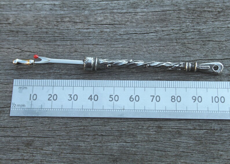 May include: A silver-colored, decorative needle with a red bead at the tip, measuring approximately 9 cm in length. The needle is designed with a twisted, textured body and a small loop at the end.