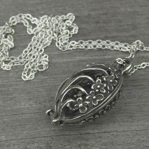 May include: A silver-colored pendant necklace with a floral design. The pendant is an oval shape with an open filigree design, showcasing small flowers. The necklace chain is also silver and lies on a gray wooden surface.