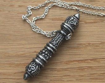 May include: A silver chain necklace with a detailed, ornate pendant shaped like a long, thin cylinder. The pendant has a textured surface with intricate designs.