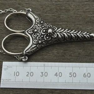 May include: Silver-toned ornate scissors with a floral design, hanging from a chain. The scissors are approximately 7 cm long.