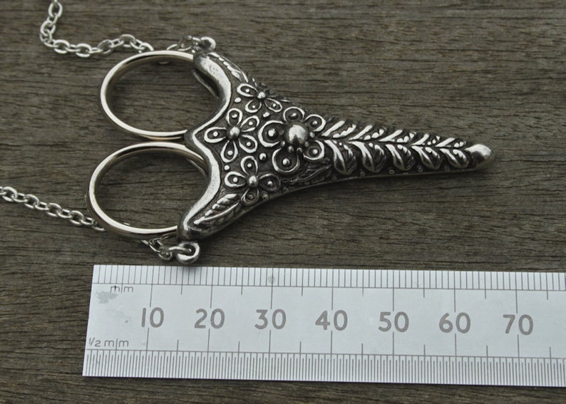 May include: Silver-toned ornate scissors with a floral design, hanging from a chain. The scissors are approximately 7 cm long.