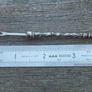 May include: A silver needle threader with a twisted handle and a red bead at the top. The needle threader is 3 inches long and is measured against a ruler.