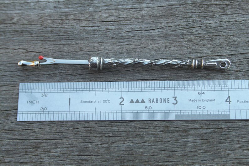 May include: A silver needle threader with a twisted handle and a red bead at the top. The needle threader is 3 inches long and is measured against a ruler.