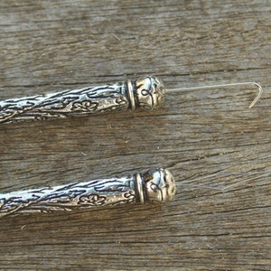 May include: Two silver-toned, ornate, decorative, wire-ended bead caps with a floral design. The bead caps are lying on a wooden surface.