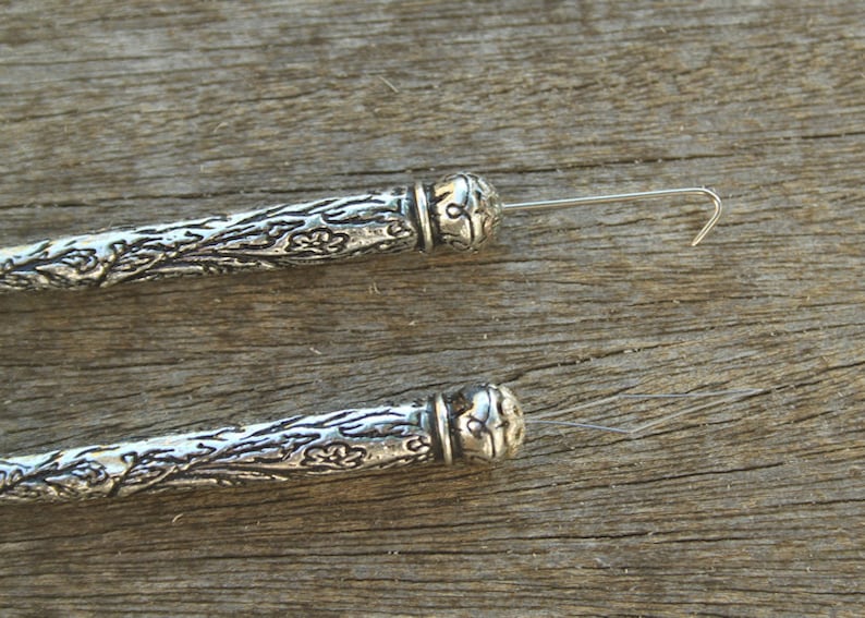 May include: Two silver-toned, ornate, decorative, wire-ended bead caps with a floral design. The bead caps are lying on a wooden surface.