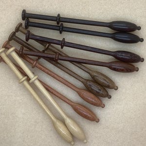 May include: A collection of wooden lace bobbins in various shades of brown and natural wood tones. The bobbins have a distinctive shape with a long, slender body and a bulbous end, used for lacemaking.