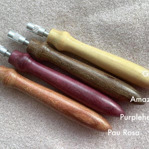 May include: Four wooden tools with metal ends, each displaying a different wood grain and colour. The wood types are labelled: Guatambu, Amazakoue, Purpleheart, and Pau Rosa. The tools are arranged diagonally on a textured surface.