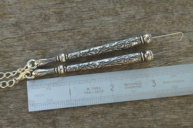 May include: Two silver needle threaders with ornate floral designs, measuring approximately 2 inches in length, are shown next to a ruler.