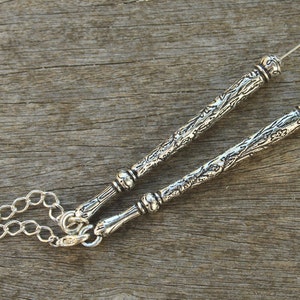 May include: A silver chain with two decorative silver rods attached. The rods have an ornate design and are connected to the chain with a loop at one end. The other end of each rod has a small loop.