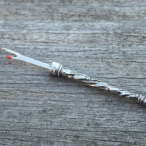 May include: A silver-colored, twisted, metal needle threader with a red tip and a small, silver-colored cap on one end.