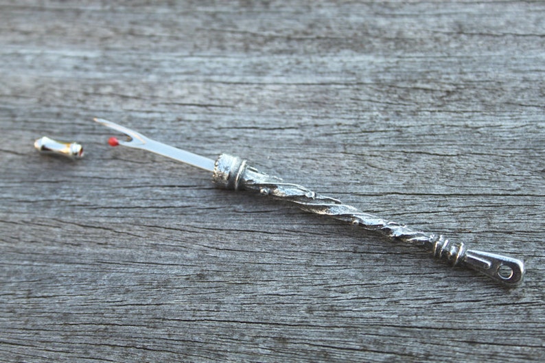 May include: A silver-colored, twisted, metal needle threader with a red tip and a small, silver-colored cap on one end.
