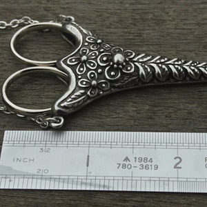 May include: Silver necklace with a pair of scissors pendant. The pendant is decorated with floral designs and has a chain attached to it. The pendant is about 2 inches long.