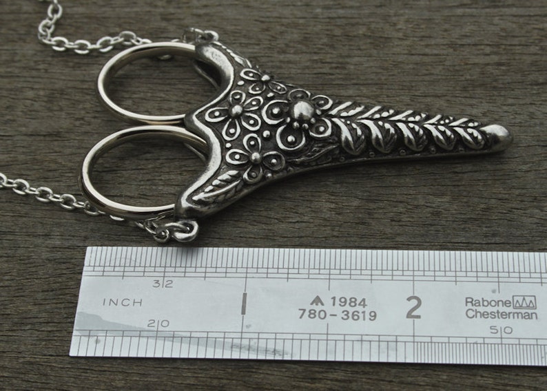 May include: Silver necklace with a pair of scissors pendant. The pendant is decorated with floral designs and has a chain attached to it. The pendant is about 2 inches long.