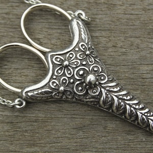 May include: A silver necklace with a pair of decorative scissors pendant. The pendant features a floral design with leaves and flowers.