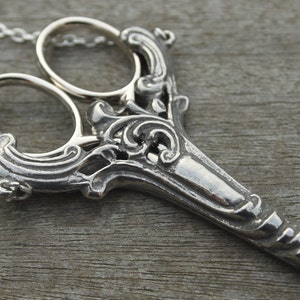 May include: A silver-colored scissor pendant with ornate detailing, suspended from a delicate chain. The scissors feature intricate scrollwork and a pointed blade, suggesting a craft or fashion theme.