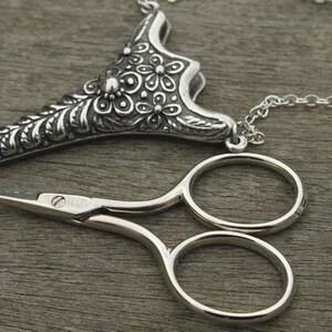 May include: A silver necklace with a floral design pendant and a pair of small silver scissors. The pendant is shaped like a teardrop and has a chain attached to it. The scissors are closed and have a silver finish.